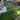 A backyard installation showing bright green artificial turf grass with a gray composite fence, concrete patio, and tree trunk in the foreground. A person's boots and feet are visible at the bottom edge of the frame.