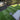 A high-angle view of a freshly installed green artificial turf lawn in a backyard, featuring a gray slatted fence on the left, a concrete patio on the right, and trees in the background. The bottom of the frame shows a person's legs and boots standing on the lawn.